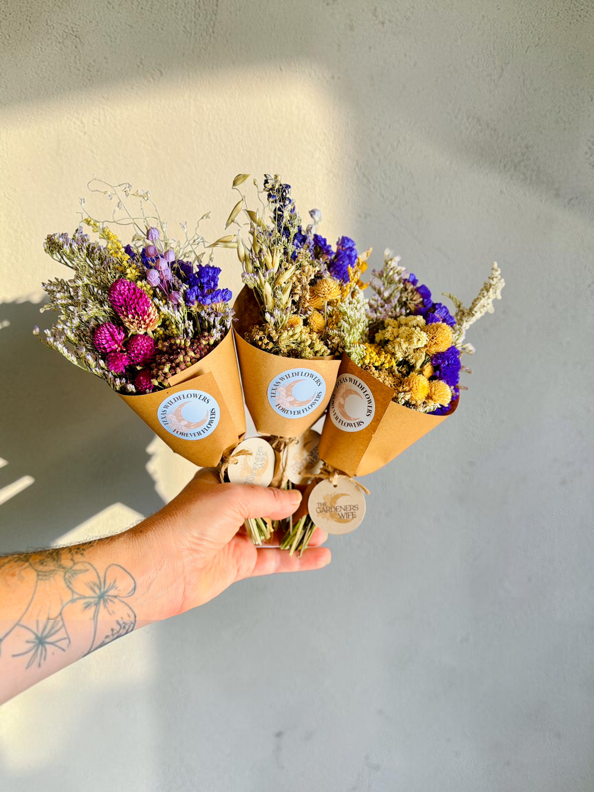Three small dried flower bouquets wrapped in brown paper cones with round blue-and-white labels, held in a person's hand against a light gray wall.