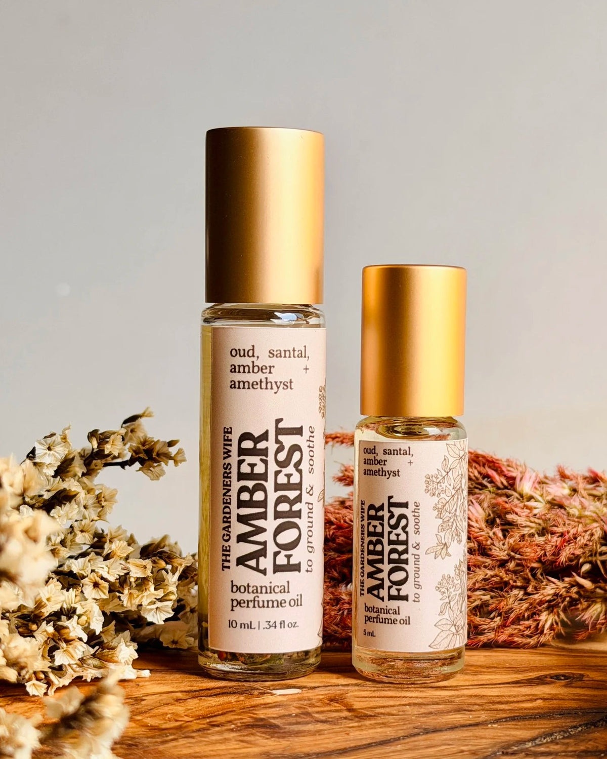 Two amber forest perfume oil roll-on bottles displayed on a rustic wooden surface with dried botanicals. One larger bottle is labeled 'oud, santal, amber,' and one smaller bottle is labeled 'amber forest botanical perfume oil'.