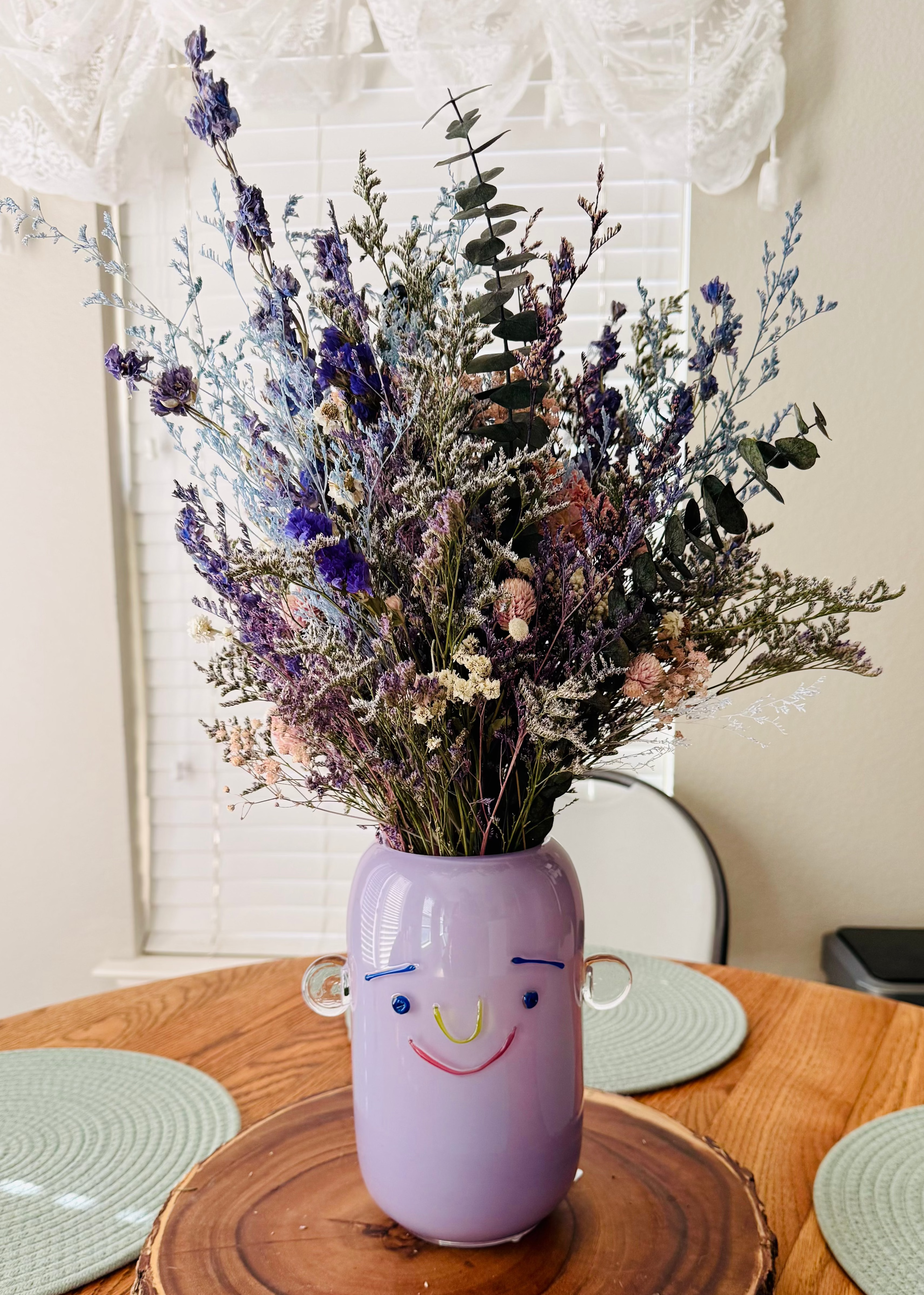 A lavender ceramic vase with a smiling face and small side handles, filled with dried purple, blue, and green flowers, displayed on a round wooden slice centerpiece on a dining table.