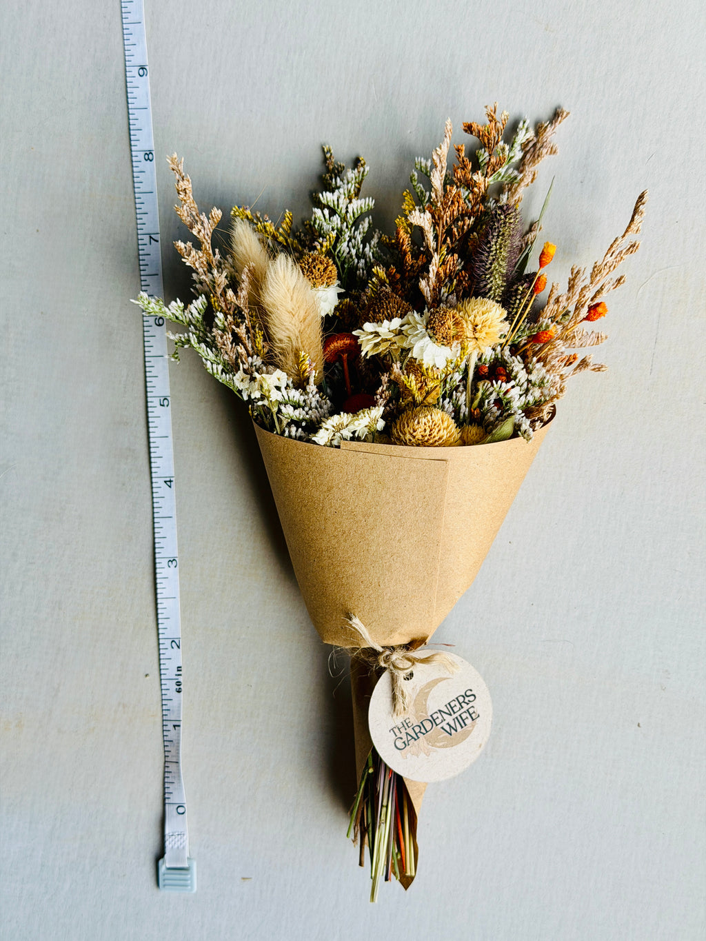 A dried flower bouquet wrapped in brown kraft paper and tied with a round tag reading 'GARDENERS WIFE', photographed against a light gray background with a measuring tape standing vertically on the left.