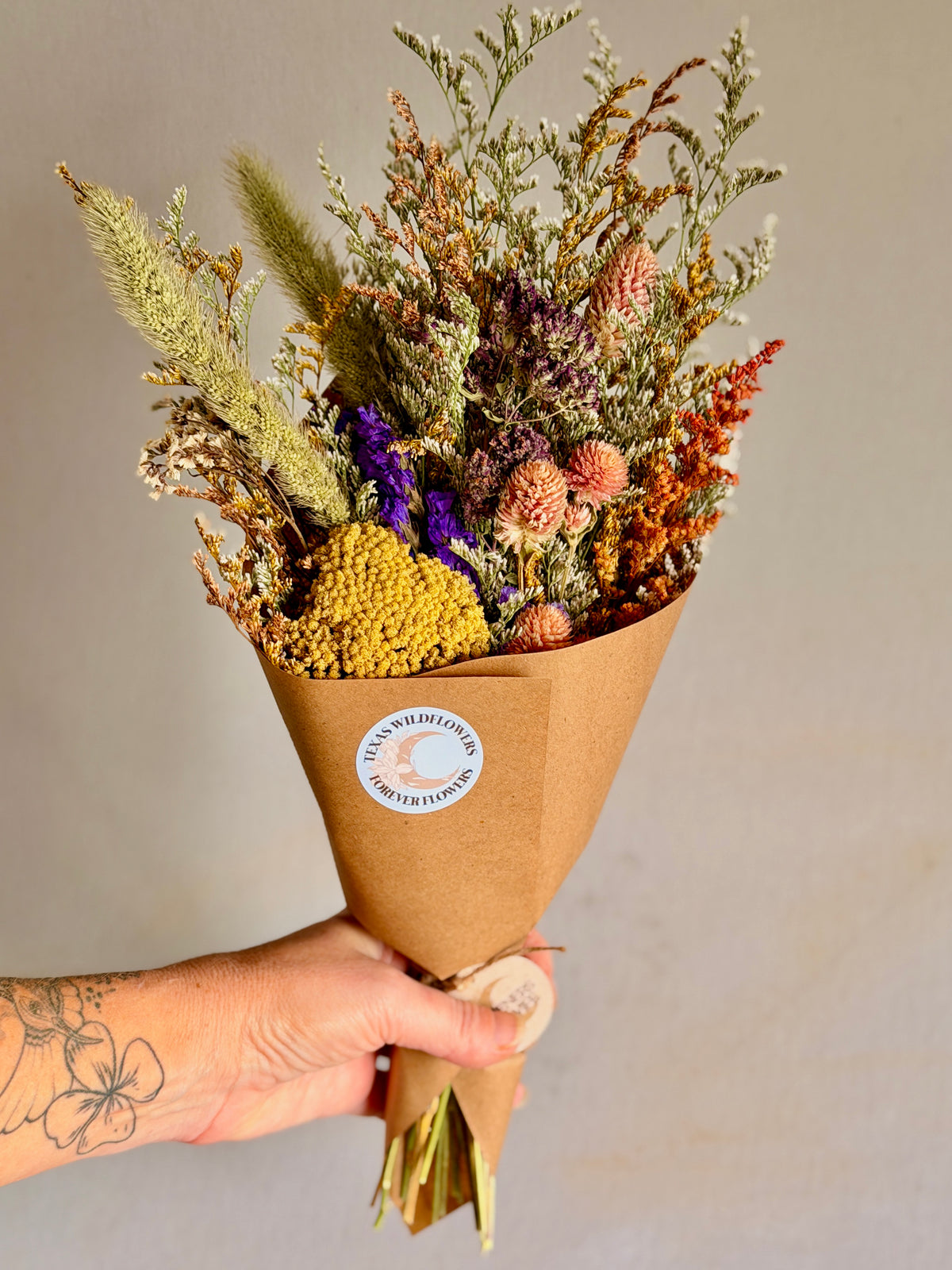 A hand holding a dried flower bouquet wrapped in brown kraft paper with a round white sticker that reads 'Texas Wildflowers Forever Flowers'. The bouquet features a variety of dried botanicals in muted shades of yellow, orange, purple, and green.