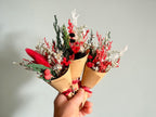 Three small dried flower bouquets wrapped in brown kraft paper cones and tied with red ribbons, held in a hand against a plain light gray background.