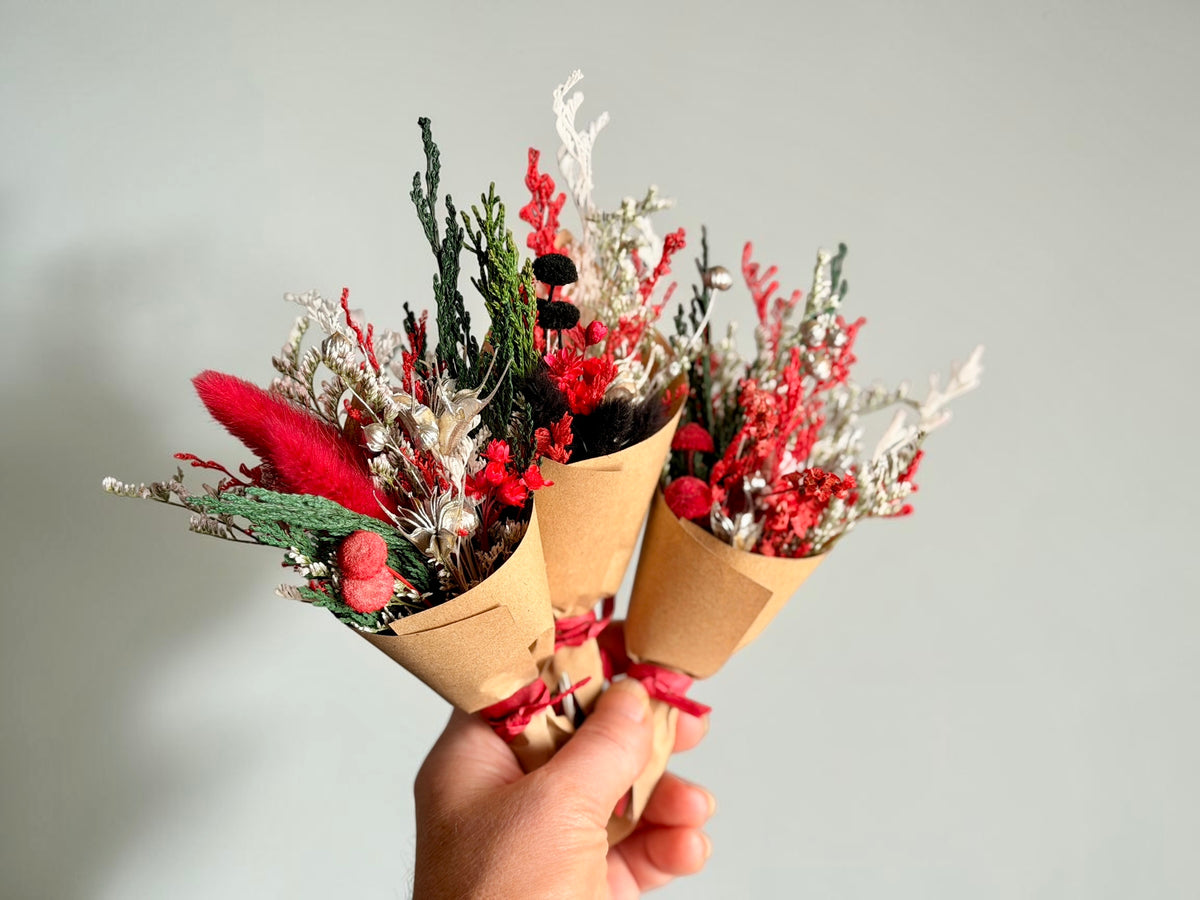 Three small dried flower bouquets wrapped in brown kraft paper cones and tied with red ribbons, held in a hand against a plain light gray background.