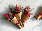 Three small dried flower bouquets wrapped in brown kraft paper and tied with red ribbons, arranged on a white marble surface. The bouquets include red berries, green pine-like stems, and pale beige dried foliage.
