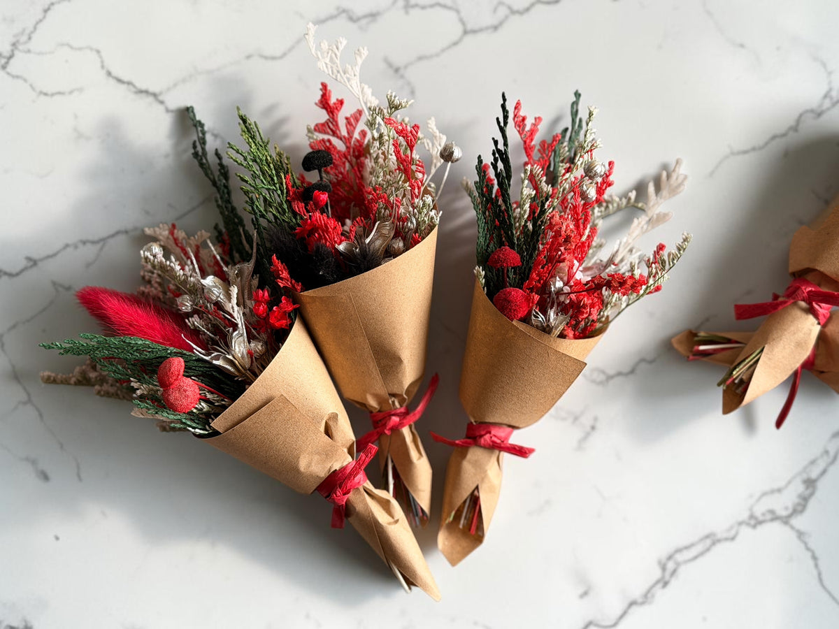 Three small dried flower bouquets wrapped in brown kraft paper and tied with red ribbons, arranged on a white marble surface. The bouquets include red berries, green pine-like stems, and pale beige dried foliage.