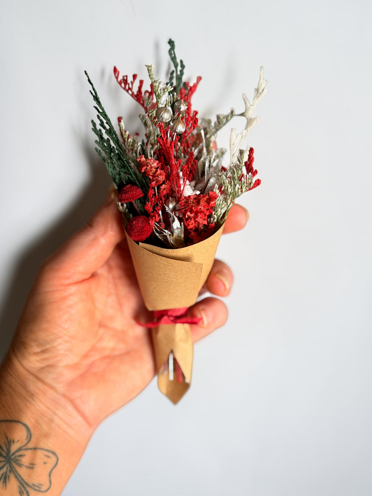A hand holding a small dried flower bouquet wrapped in brown kraft paper and tied with a red ribbon, against a plain light gray background.