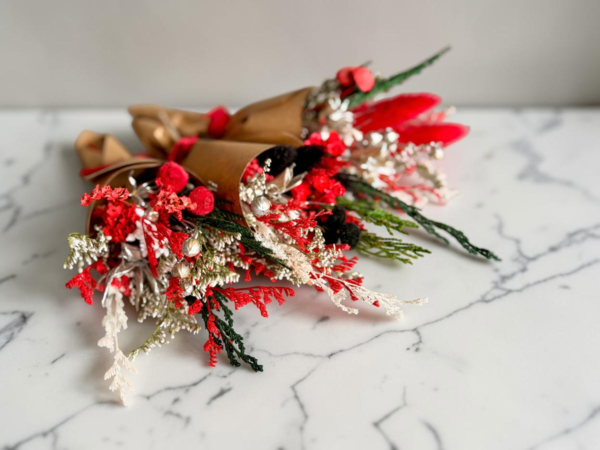three small dried flower bouquets wrapped in brown paper, featuring vibrant red blossoms, white textured filler, and green pine needles, arranged on a white marble surface.