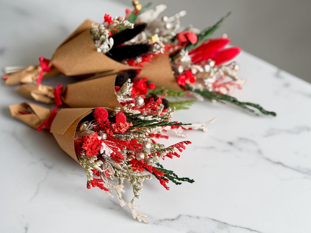 Three small dried flower bouquets wrapped in brown kraft paper and tied with red ribbons, arranged on a white marble surface. The bouquets include red dried flowers, silver glittery filler, and green stems.