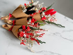 Three small dried flower bouquets wrapped in brown kraft paper and tied with red ribbons, arranged on a white marble surface. The bouquets include red dried flowers, silver glittery filler, and green stems.
