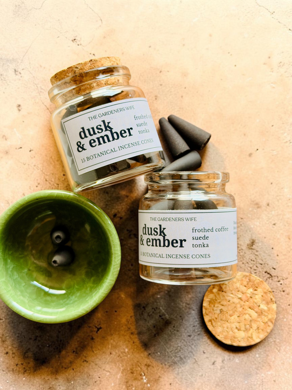 Two glass jars of 'dusk & ember' botanical incense cones by The Gardeners Wife, styled on a textured beige stone surface with a green ceramic bowl, wooden wicks, and a cork.