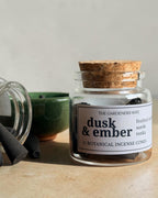 A clear glass jar with a cork stopper and a white label reading 'dusk & ember' sits on a stone surface, with several dark incense cones beside it and another jar lying on its side in the background.