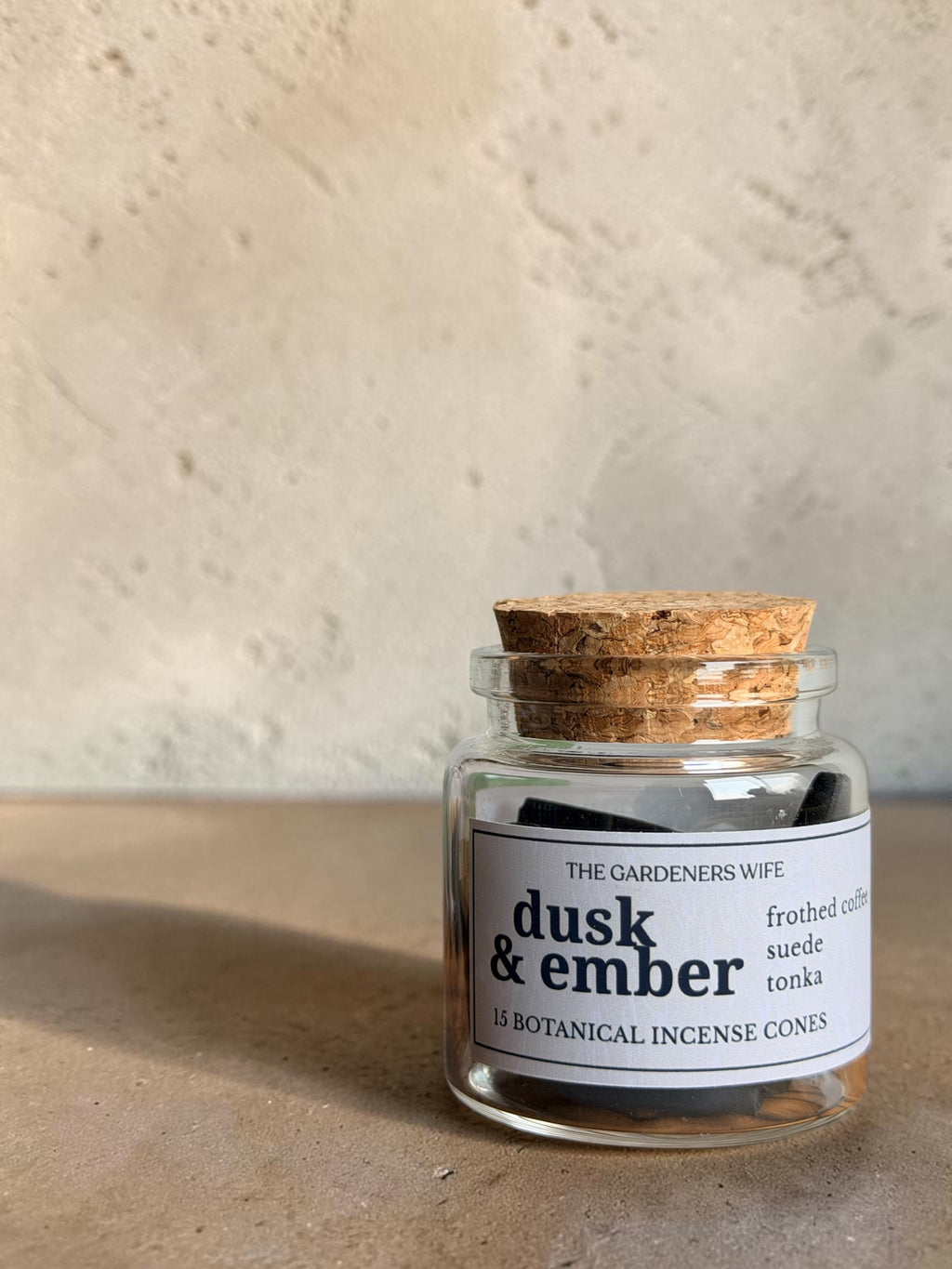 A small clear glass jar candle with a cork stopper and a white label reading 'dusk & ember' by The Gardeners Wife, set on a textured beige surface against a matching beige wall.