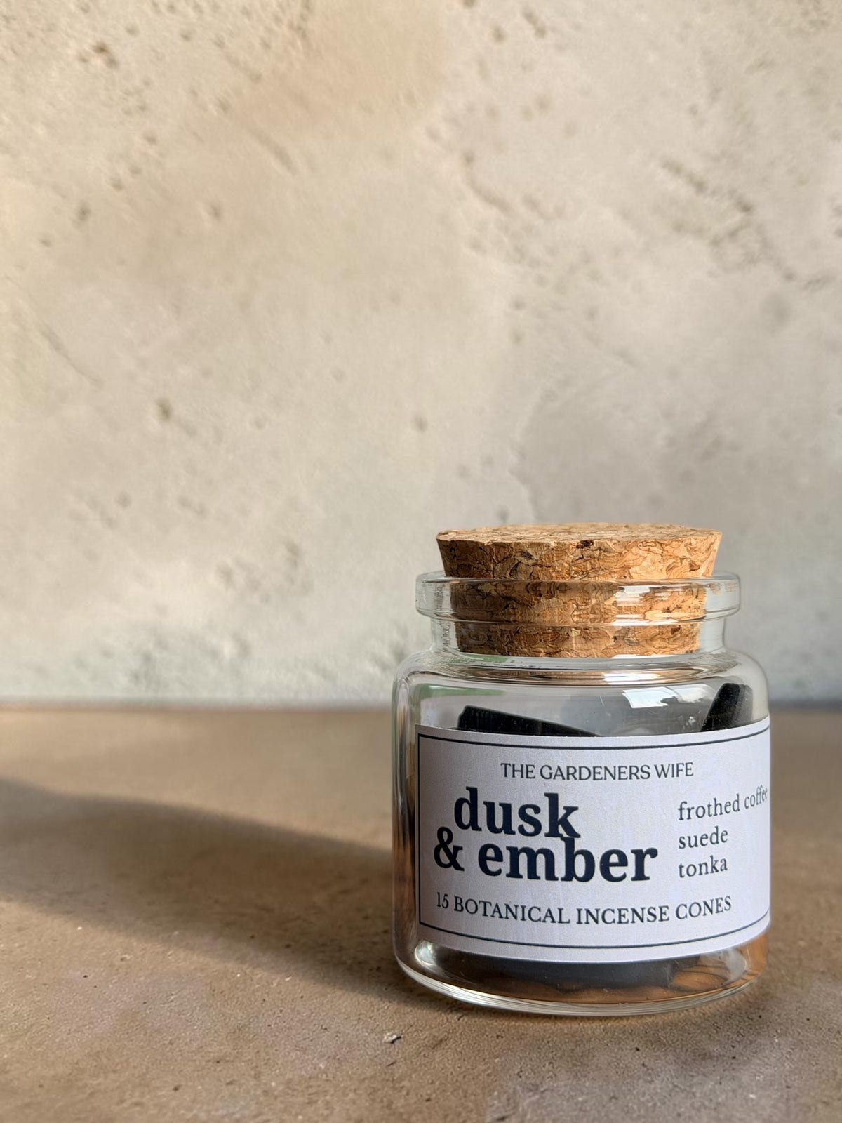 A small clear glass jar candle with a cork stopper and a white label reading 'dusk & ember' by The Gardeners Wife, set on a textured beige surface against a matching beige wall.
