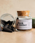A glass jar candle labeled 'dusk & ember' with a cork stopper, shown beside several dark incense cones and a cut cork on a beige surface, with a green bowl partially visible in the background.