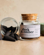 A glass jar candle labeled 'dusk & ember' with a cork stopper, shown beside several dark incense cones and a cut cork on a beige surface, with a green bowl partially visible in the background.