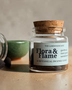 A clear glass jar with a cork lid and a white label reading 'Flora & Flame' by The Gardeners Wipe, shown on a beige surface with a few dark incense cones beside an tipped-over jar and a green bowl in the background.