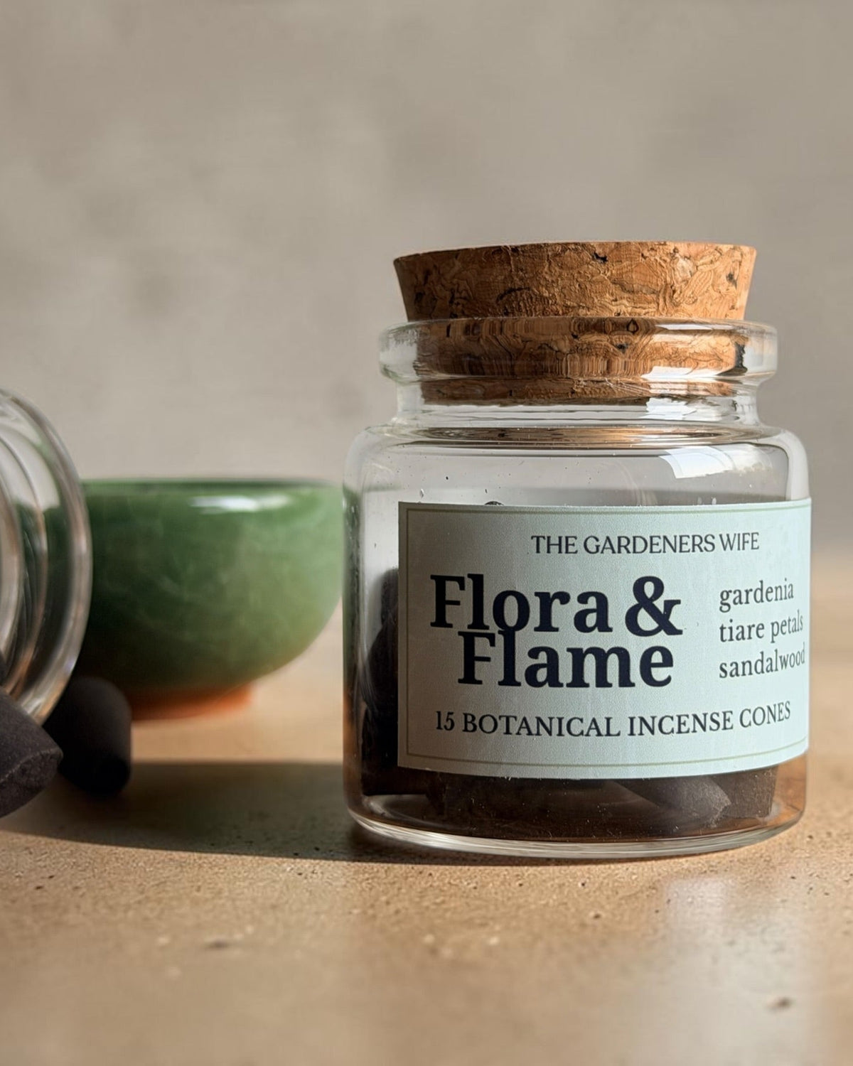 A clear glass jar with a cork lid and a white label reading 'Flora & Flame' by The Gardeners Wipe, shown on a beige surface with a few dark incense cones beside an tipped-over jar and a green bowl in the background.