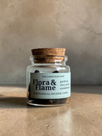 A small clear glass jar with a cork stopper and a white label reading 'Flora & Flame' by The Gardeners Wife, placed on a textured beige surface against a neutral beige wall.