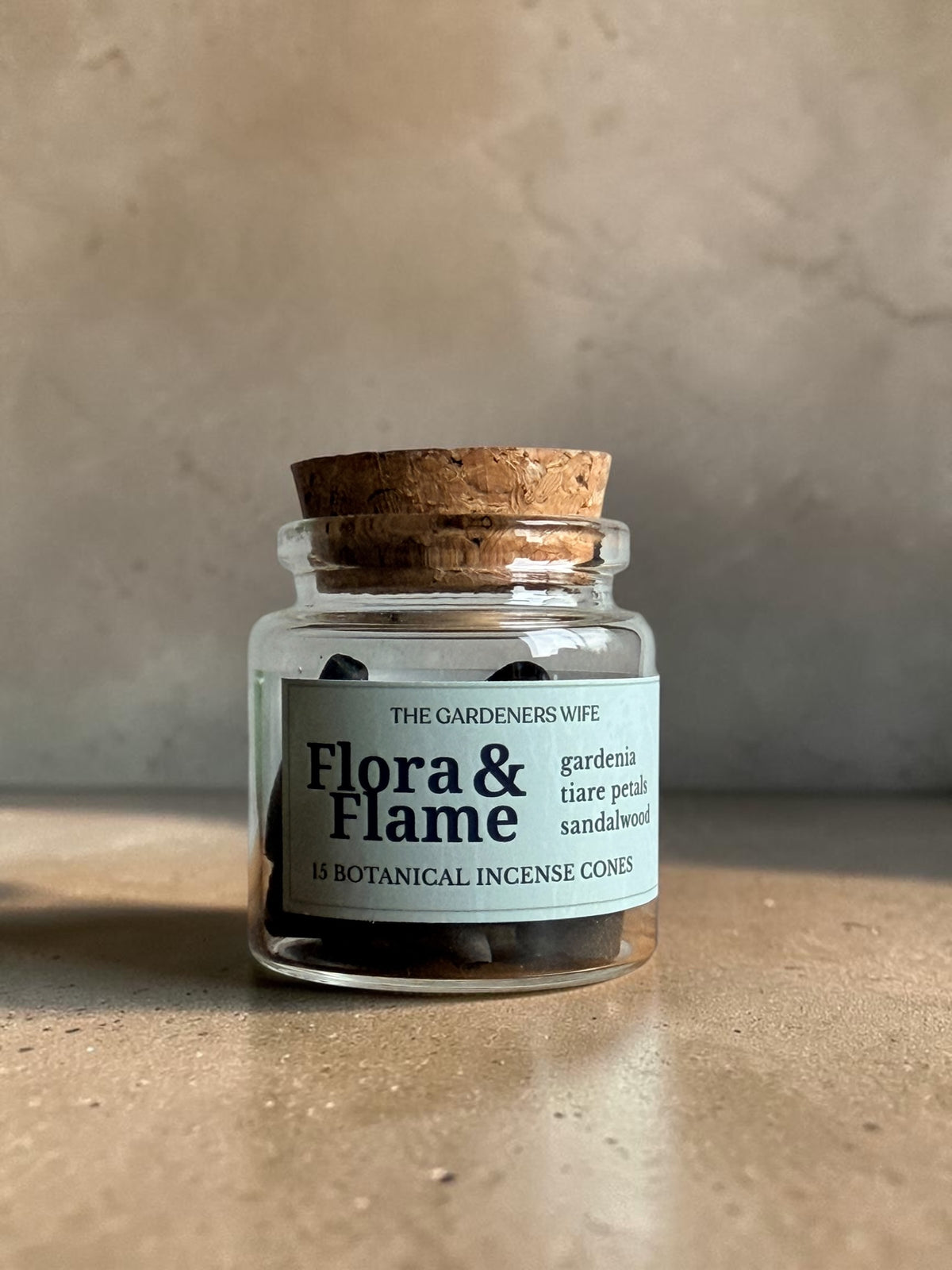 A small clear glass jar with a cork stopper and a white label reading 'Flora & Flame' by The Gardeners Wife, placed on a textured beige surface against a neutral beige wall.