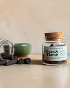 A small clear glass jar with a cork stopper and a white label reading 'Flora & Flame' sits on a beige surface. A second jar lies on its side with dark incense sticks spilled out, along with a green bowl  in the background.