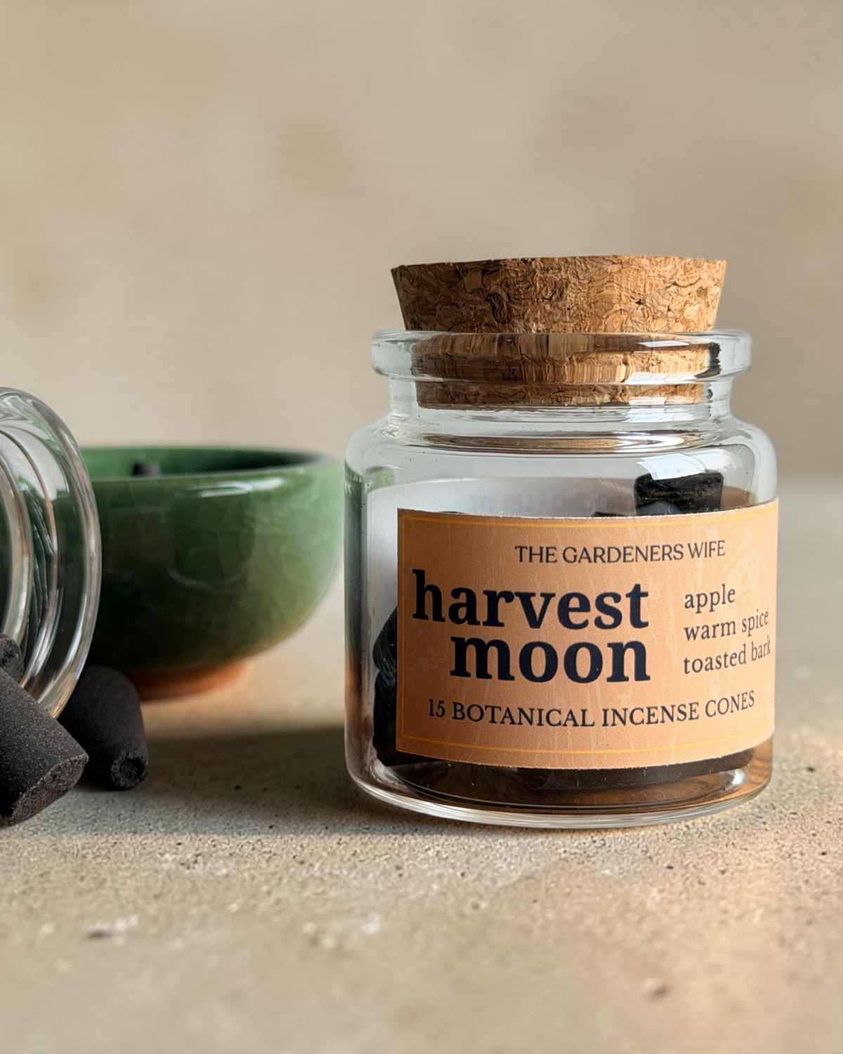 A small clear glass jar with a cork stopper and a tan label reading 'harvest moon' stands on a beige surface. A second jar lies on its side with loose dark incense cones spilled out, and a small green bowl is behind them.