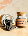 Two small glass jars with cork lids on a stone surface, one lying on its side with dark incense cones spilling out and one standing upright with an orange label reading 'harvest moon'.