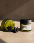 A small glass jar candle with a black lid and white label sits beside an overturned matching jar filled with dark incense cones on a beige stone surface in warm sunlight.