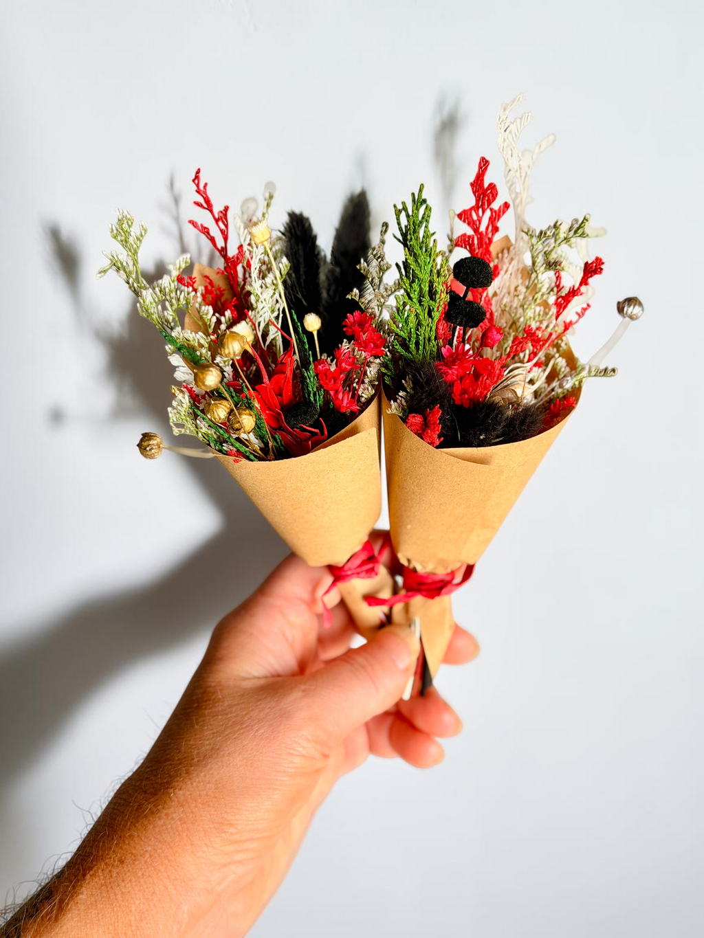 A hand holding two small dried flower bouquets wrapped in brown kraft paper cones with red ribbon, set against a plain light gray background.