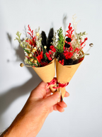 A hand holding two small dried flower bouquets wrapped in brown kraft paper cones with red ribbon, set against a plain light gray background.