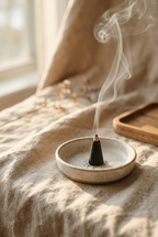 A small round ceramic incense holder with a smoking incense cone, set on a draped beige linen surface with soft sunlight and a wooden tray in the background.