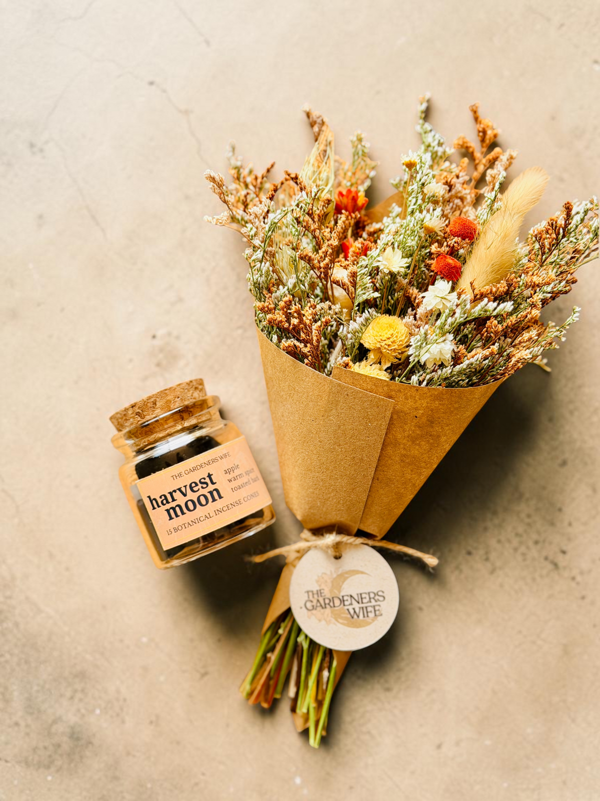 A small glass jar labeled 'harvest moon' sits beside a wrapped bouquet of dried flowers in brown kraft paper with a round tag reading 'The Gardeners Wife,' on a light beige textured surface.