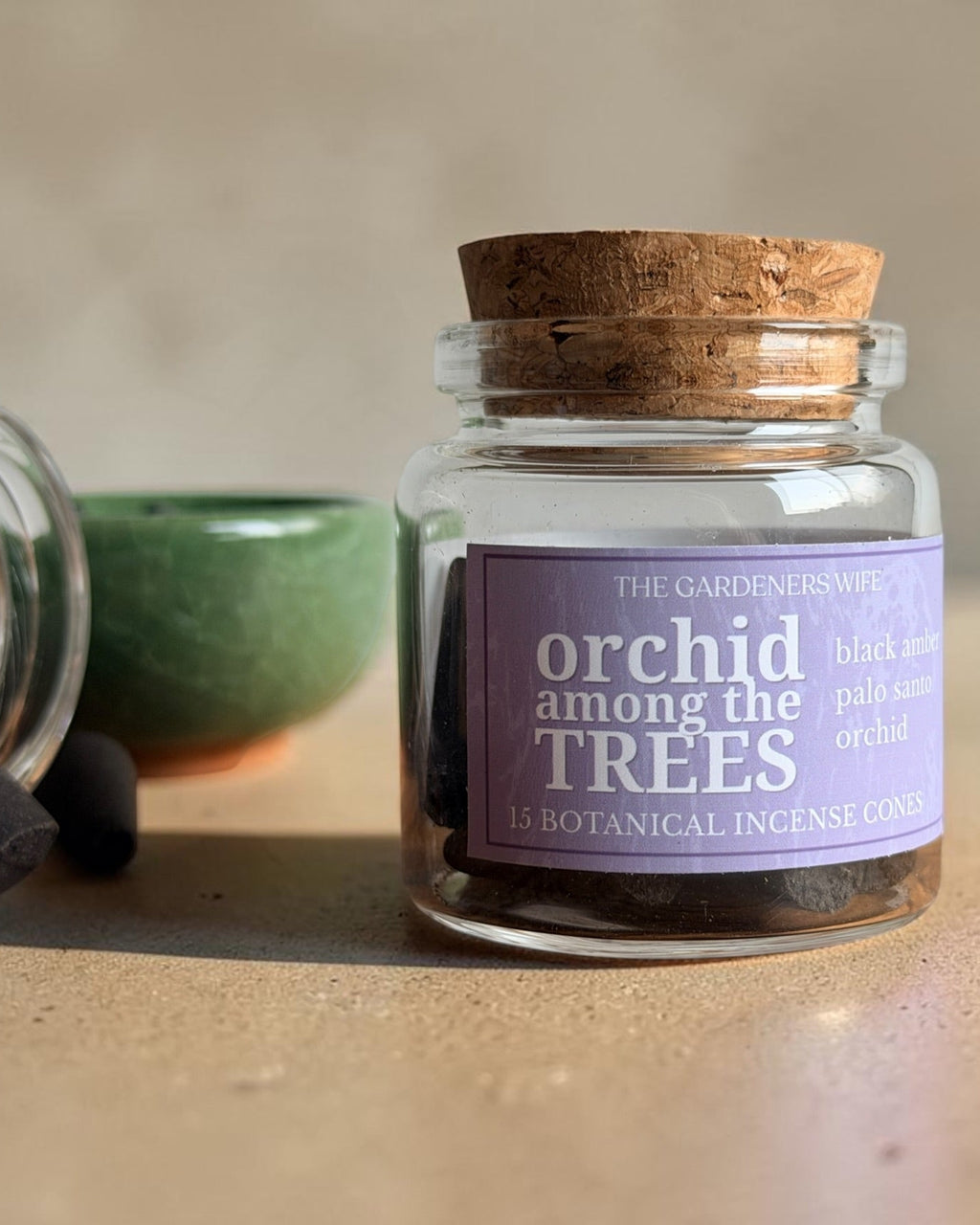 A small clear glass jar with a cork stopper and a lavender label reading 'orchid among the TREES' by The Gardener's Wife, shown on a beige surface with incense cones and a green bowl nearby.