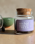 A small clear glass jar with a cork stopper and a lavender label reading 'orchid among the TREES' by The Gardener's Wife, shown on a beige surface with incense cones and a green bowl nearby.