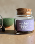 A small clear glass jar with a cork stopper and a lavender label reading 'orchid among the TREES' by The Gardener's Wife, shown on a beige surface with incense cones and a green bowl nearby.