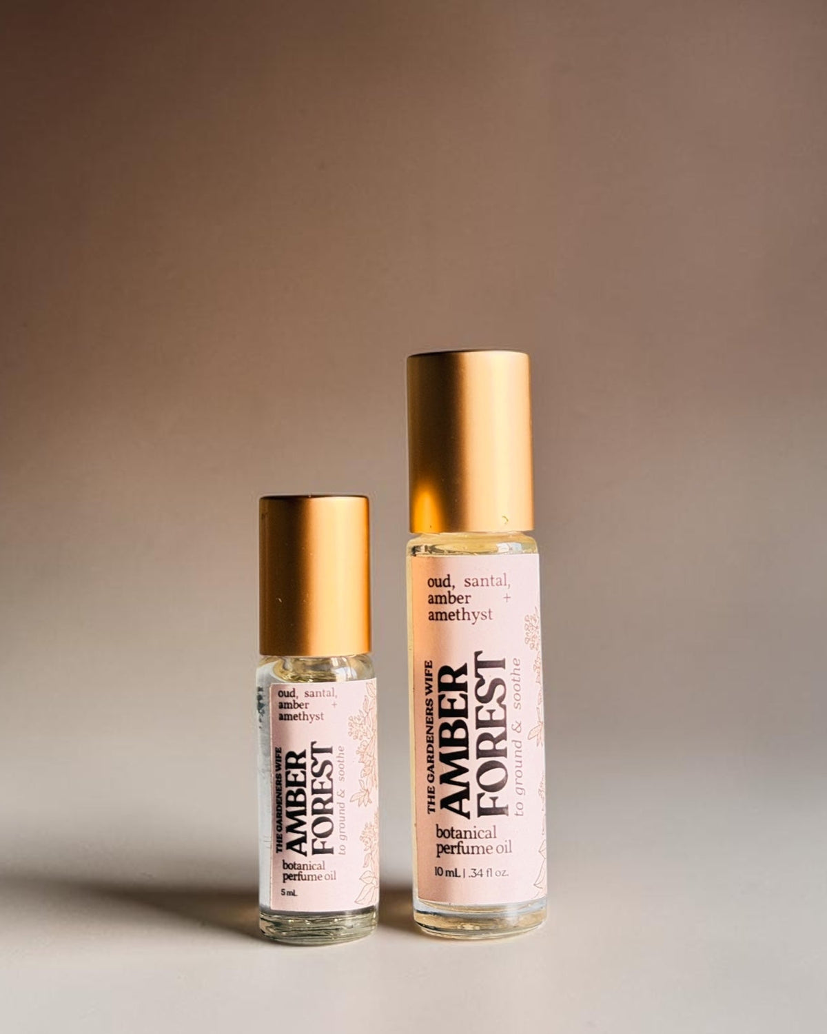 Two amber forest roll-on bottles with gold caps and cream labels, standing on a light surface against a warm brown gradient background.