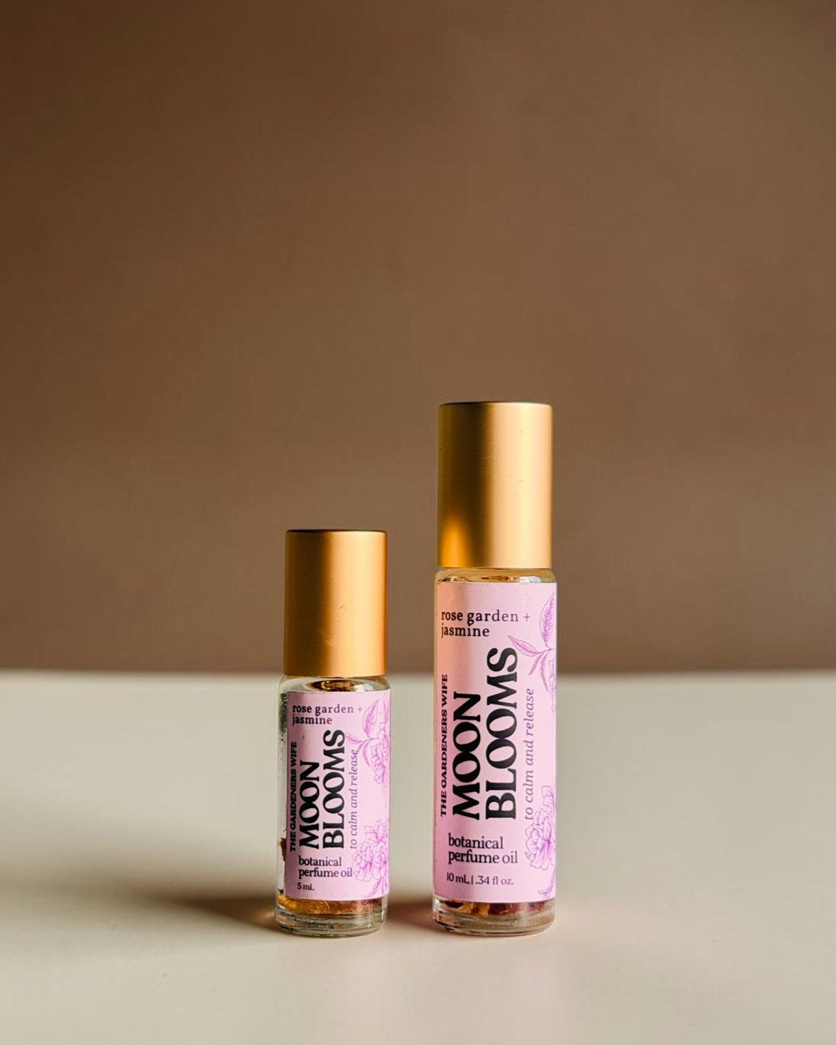 Two small cosmetic roller bottles labeled 'MOON BLOOMS' with pink labels and gold caps, standing on a light surface against a warm brown background.