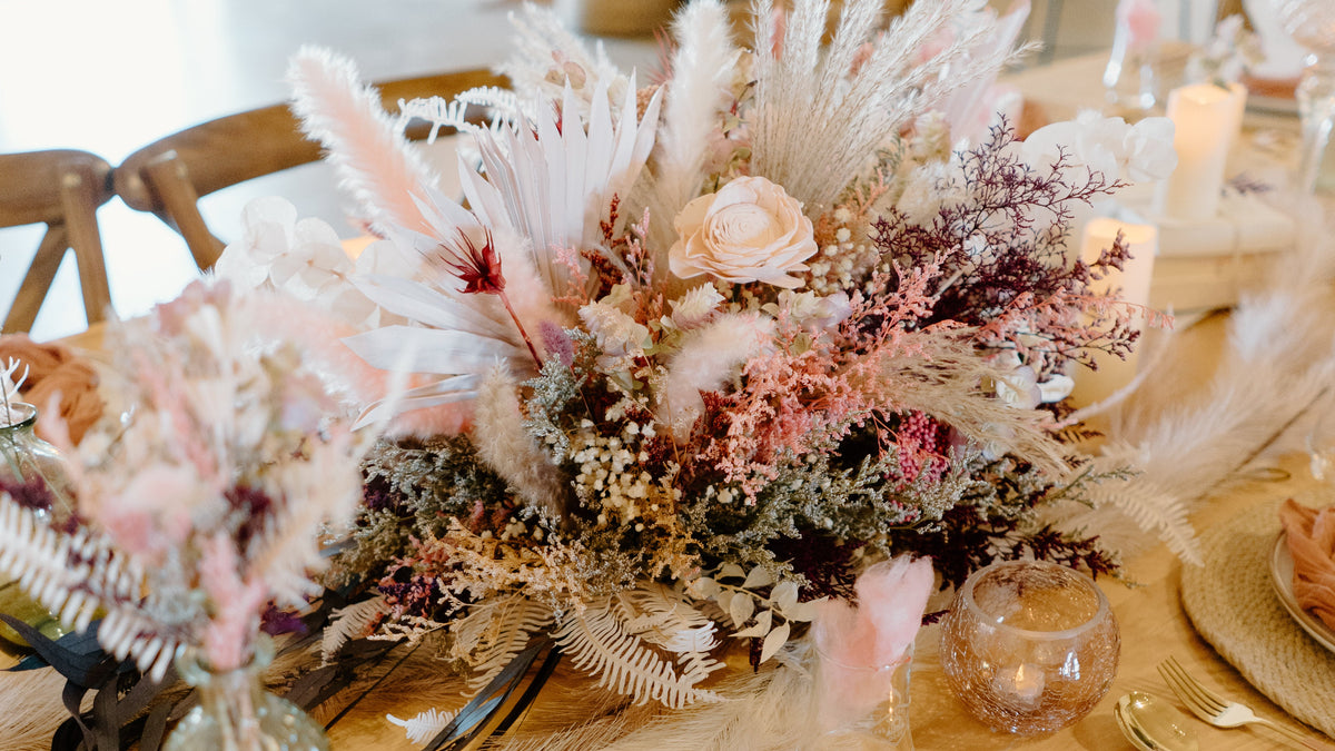 A styled wedding table setting featuring a large central dried floral arrangement with pink, white, and dusty purple botanicals, a textured gold vase, woven placemats, white plates, blush napkins, and gold cutlery in a bright, airy event space.